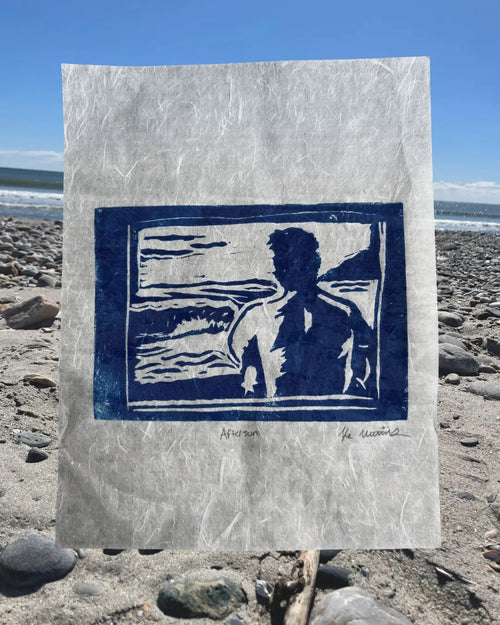 A square art print with a textured off-white paper background and a dark blue linocut-style illustration of a figure by the sea, photographed standing upright on a rocky beach with the ocean and blue sky in the background.