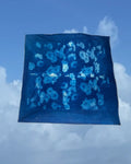 A square blue textile with a white floral cyanotype pattern, photographed floating against a blue sky with clouds.